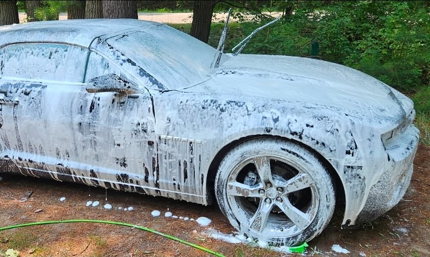 Thick white soap foam covers a car parked outdoors with its windshield wipers raised.
